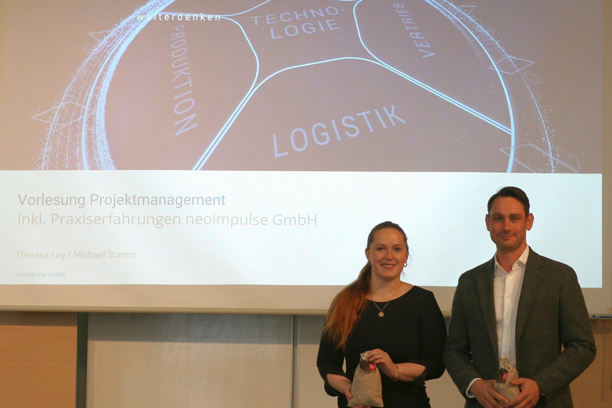 Theresa Loy und Michael Stamm, beide neoimpulse GmbH, stehen in der Aula vor einer Leinwand, auf der die PowerPoint-Präsentation "Vorlesung Projektmanagement inkl. Praxiserfahrungen neoimpulse GmbH" dargestellt wird.