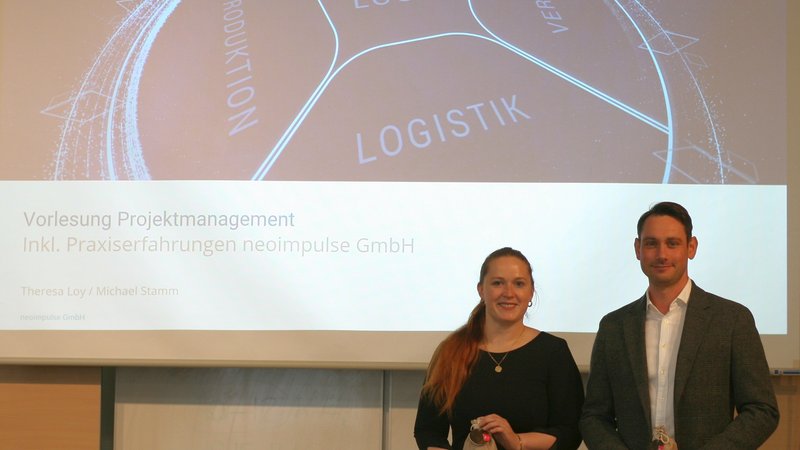 Theresa Loy und Michael Stamm, beide neoimpulse GmbH, stehen in der Aula vor einer Leinwand, auf der die PowerPoint-Präsentation "Vorlesung Projektmanagement inkl. Praxiserfahrungen neoimpulse GmbH" dargestellt wird.