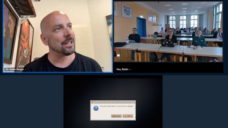 Links oben wird ein Fenster mit dem zugeschalteten Herrn Dr. Bargas gezeigt, rechts oben ein Fenster mit den Teilnehmenden in der Aula. Unten findet sich ein Fenster in der Mitte mit dem Text: "Do you really want to cancel the upload? Cancel/OK".