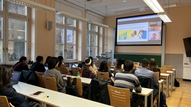 Das Bild zeigt Studenten des Studiengangs "International Business and Economics“ im Hörsaal, die dem Online-Vortrag von Ricardo Aceves auf der Projektionsleinwand folgen.