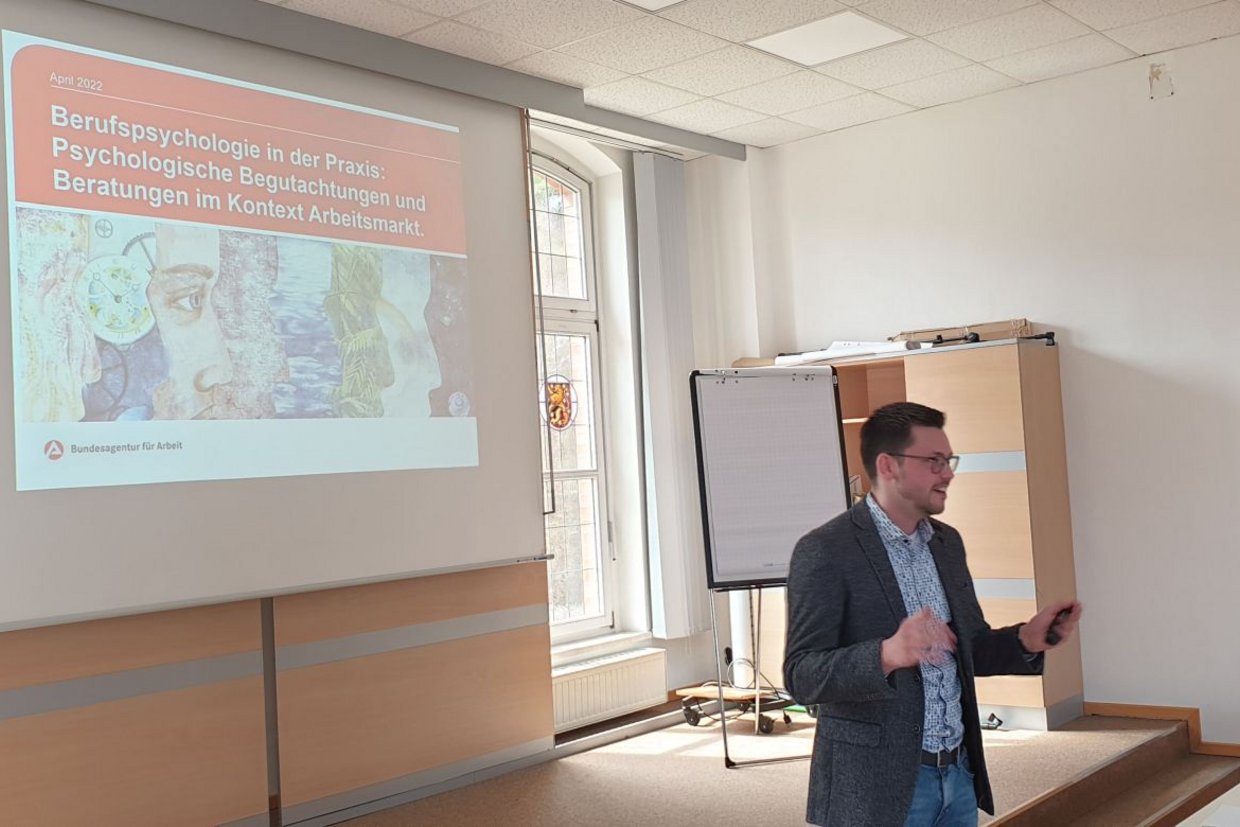 Das Bild zeigt den Psychologen Herr Lubensky in der Aula beim Vortrag. Im Hintergrund ist die Eröffnungsfolie mit der Überschrift "Berufspsychologie in der Praxis: Psychologische Begutachtungen und Beratungen im Kontext Arbeitsmarkt" erkennbar.