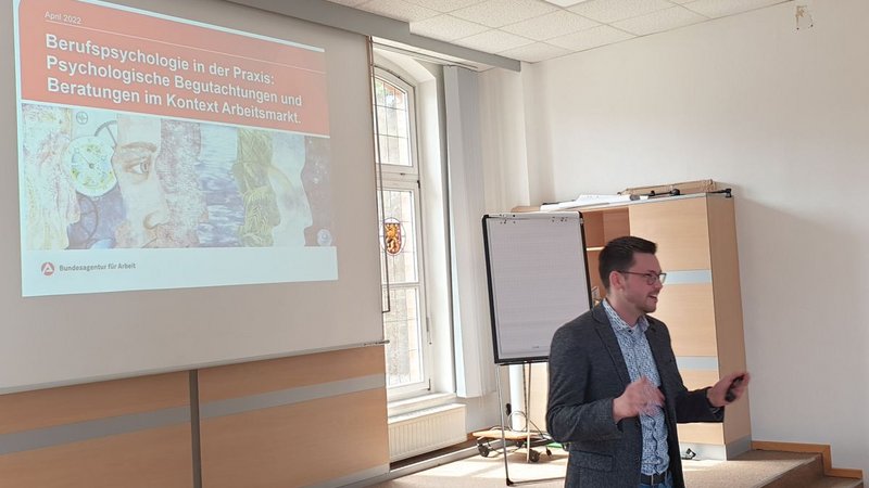 Das Bild zeigt den Psychologen Herr Lubensky in der Aula beim Vortrag. Im Hintergrund ist die Eröffnungsfolie mit der Überschrift "Berufspsychologie in der Praxis: Psychologische Begutachtungen und Beratungen im Kontext Arbeitsmarkt" erkennbar.