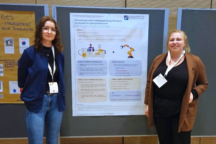 Emely Weber (links) und Maja Böhme (rechts) stehen vor ihrem Poster auf dem Herbstkongress Zwei Studentinnen (Emely Weber und Maja Böhme) in formeller Kleidung stehen vor einem großen Poster. Das Poster hängt an einer Pinnwand. Es ist zu weit weg, um den Text darauf lesen zu können.
