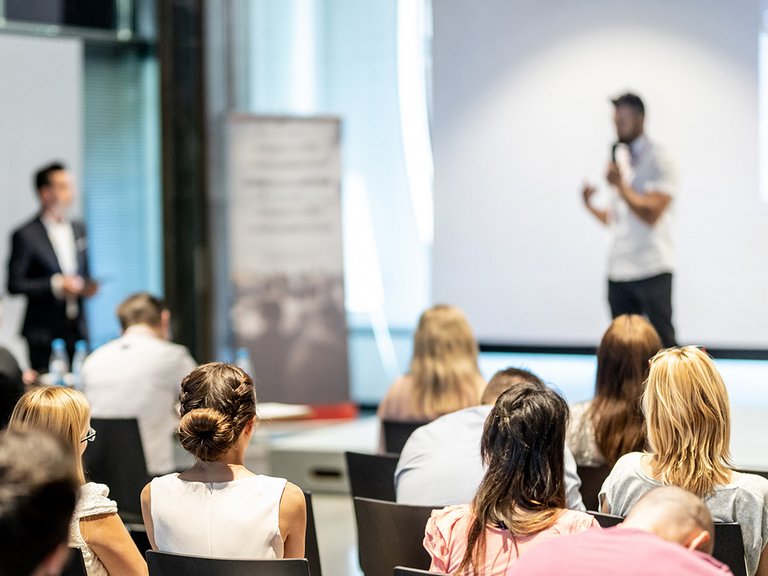 Menschen in einem Raum sitzend vor einer Bühne mit einem Speaker