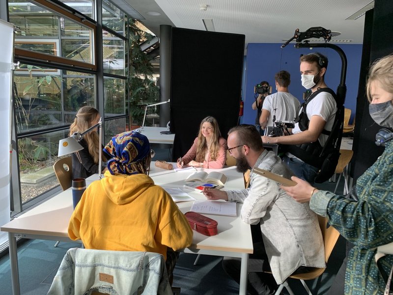 3) Drehszene in der Bibliothek: vier Studierende in Kleingruppenarbeit, das Drehteam nimmt auf.